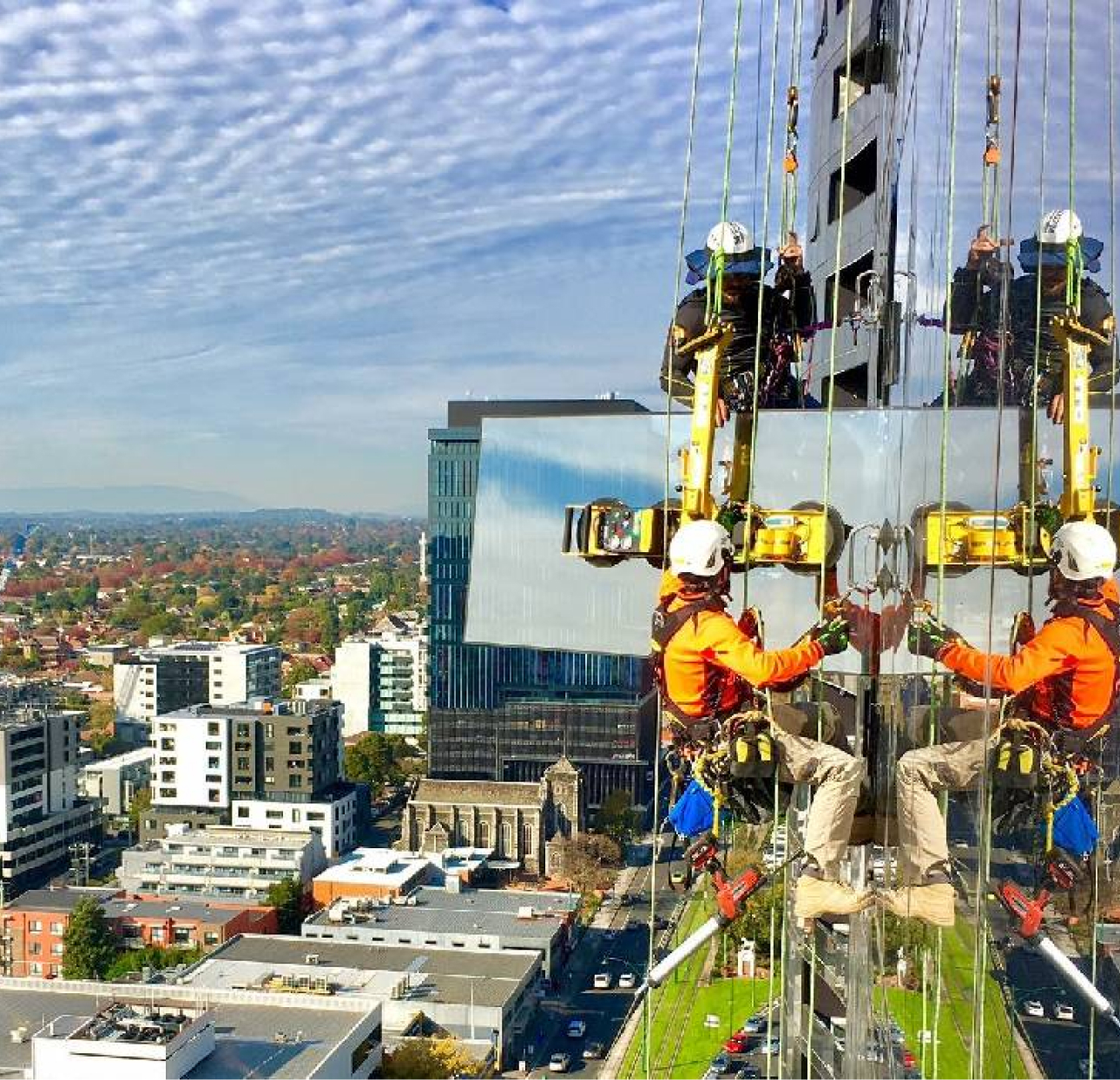 Construction workers are installing large glass panels on a high-rise building with a city skyline in the background.