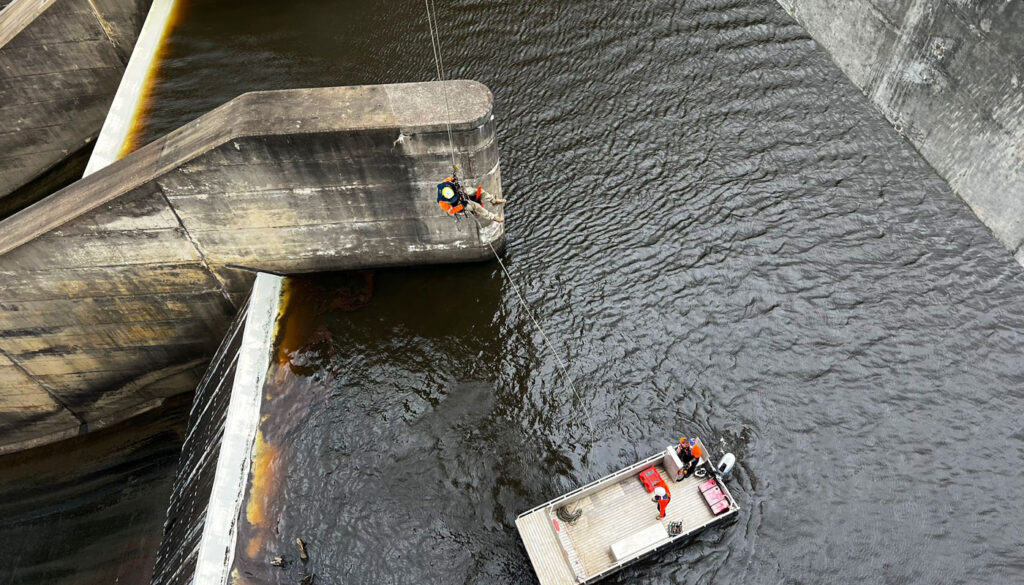 Workers inspect a dam structure from a boat and while suspended by ropes.
