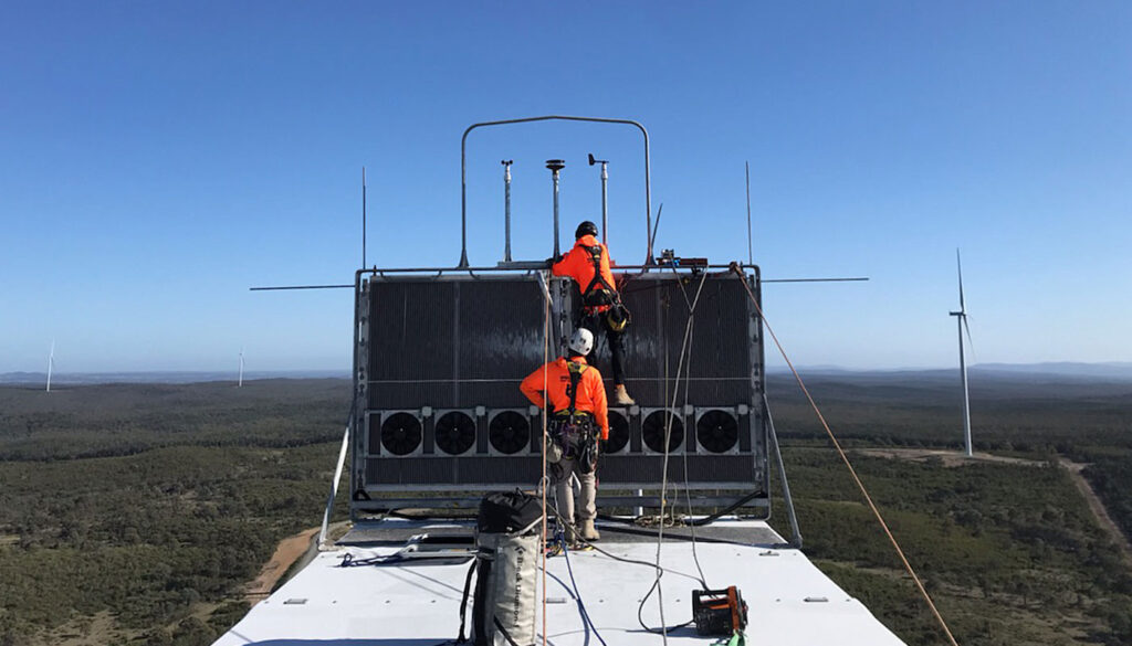Two workers in safety gear are inspecting equipment on top of a structure with wind turbines visible in the background.
