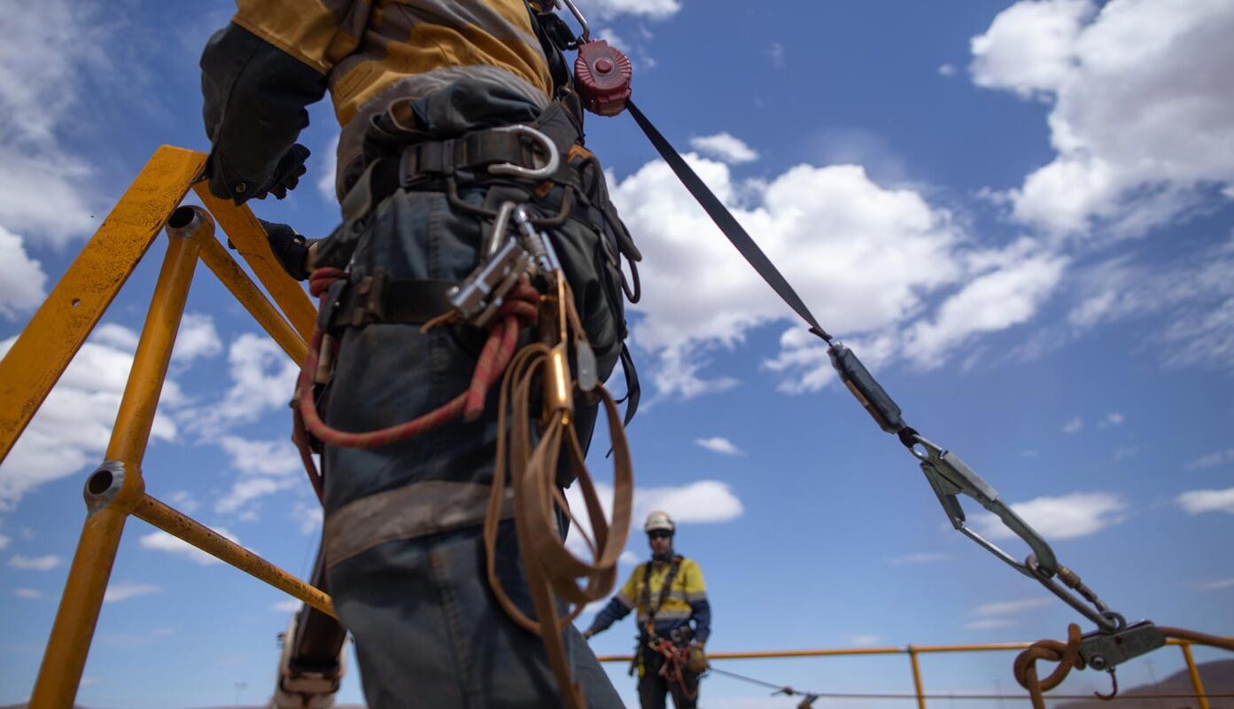 A construction worker wearing a safety harness is secured to a cable while working at height with a colleague in the background.