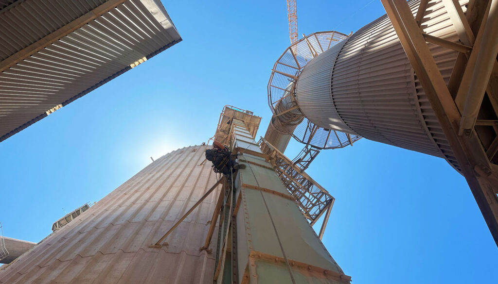 A low-angle view of industrial structures with a worker rappelling down a tall silo under a clear blue sky.