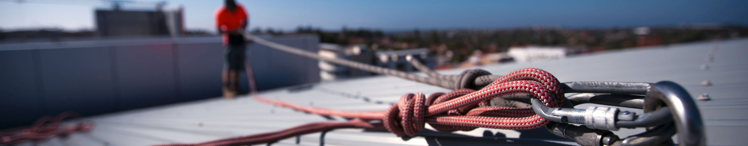 A person stands on a rooftop with a safety rope and harness in the foreground.
