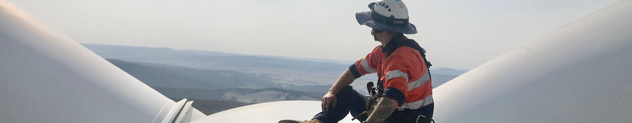 A worker wearing a helmet and safety gear sits on a wind turbine blade with a landscape view in the background.