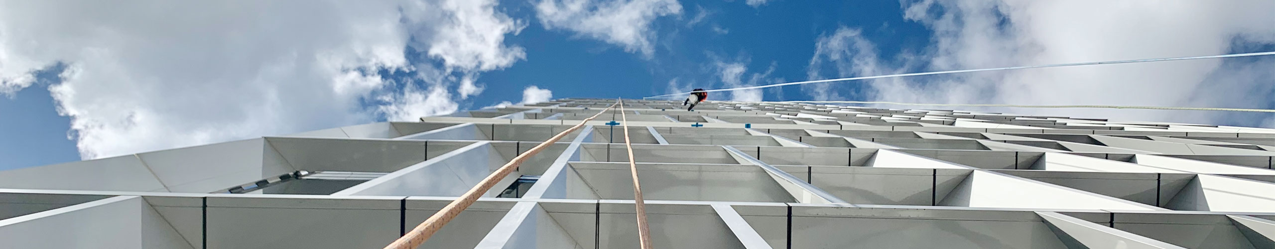 A worms-eye view of a modern building with a person rappelling down its facade against a cloudy blue sky.