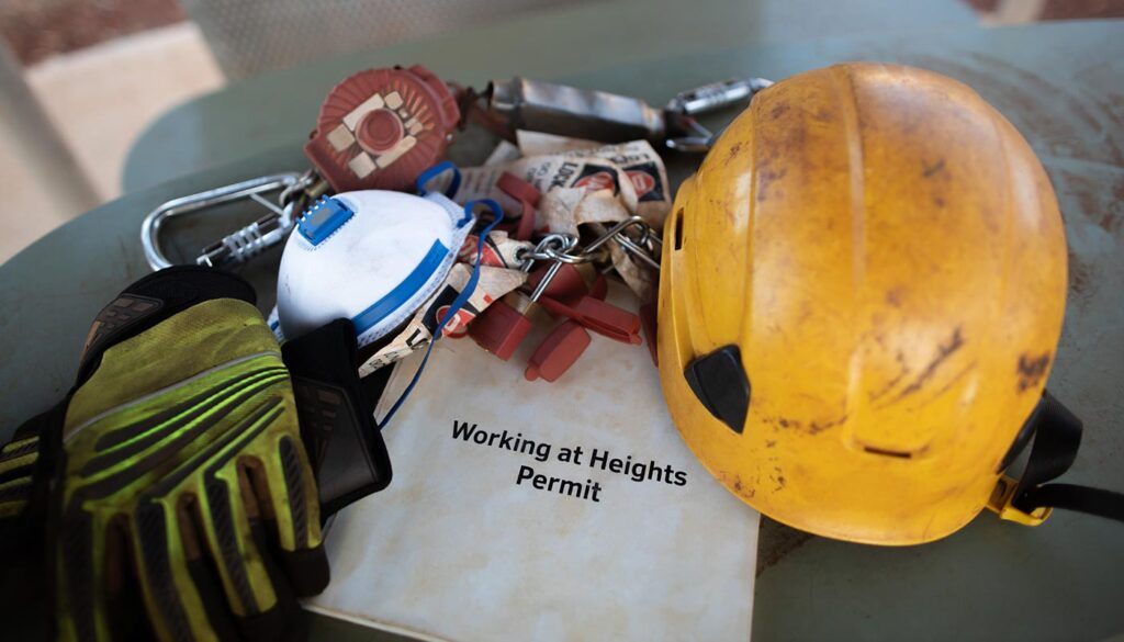 Personal protective equipment (PPE) including a hard hat, gloves, respirator, and a Working at Heights Permit are arranged on a metal surface.