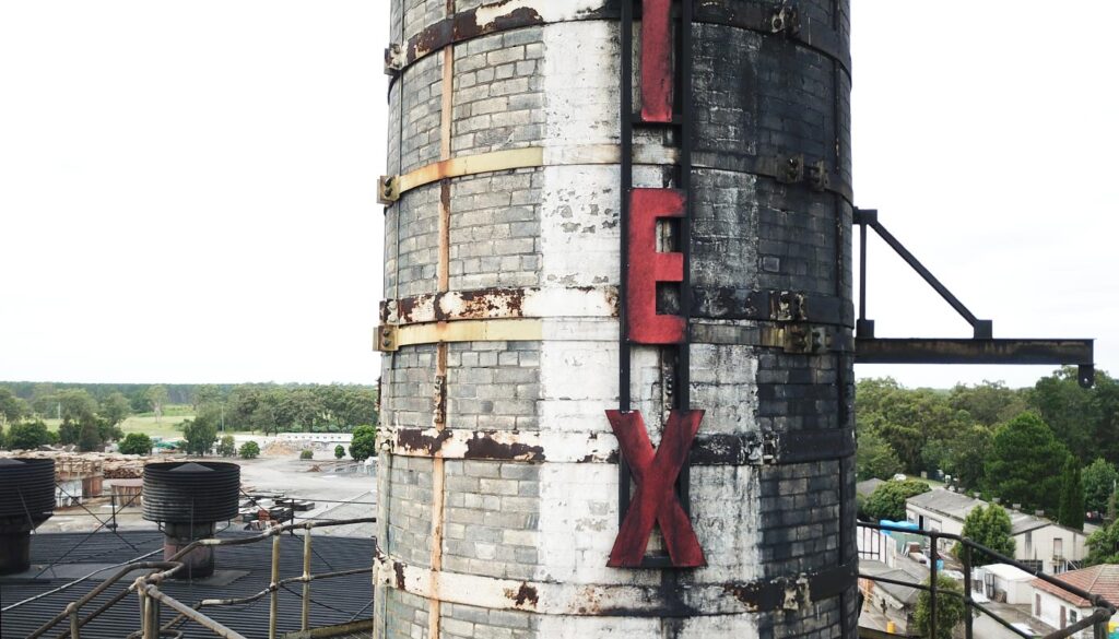 A tall, weathered industrial chimney with a large red X painted on its side, overlooking a landscape of trees and buildings.