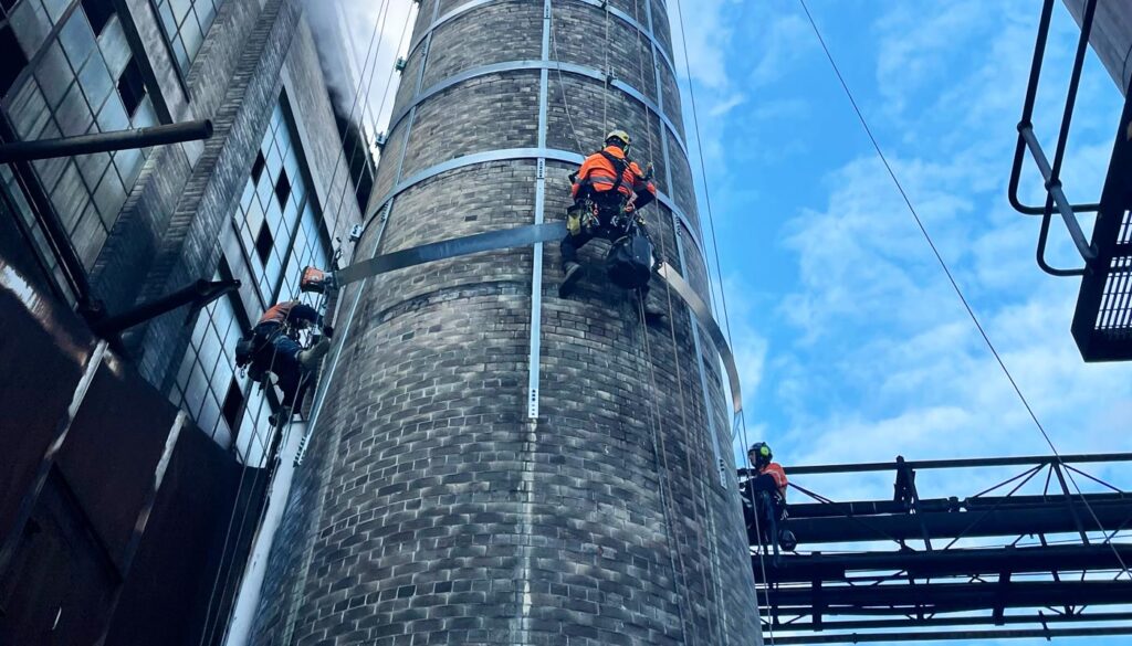 Workers are suspended on ropes while inspecting or maintaining a tall industrial chimney under a blue sky.