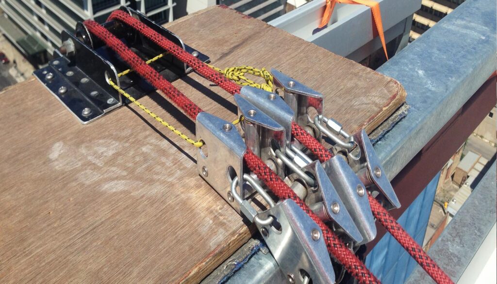 A close-up of a fall protection system with ropes and clamps secured to a wooden platform on a construction site.