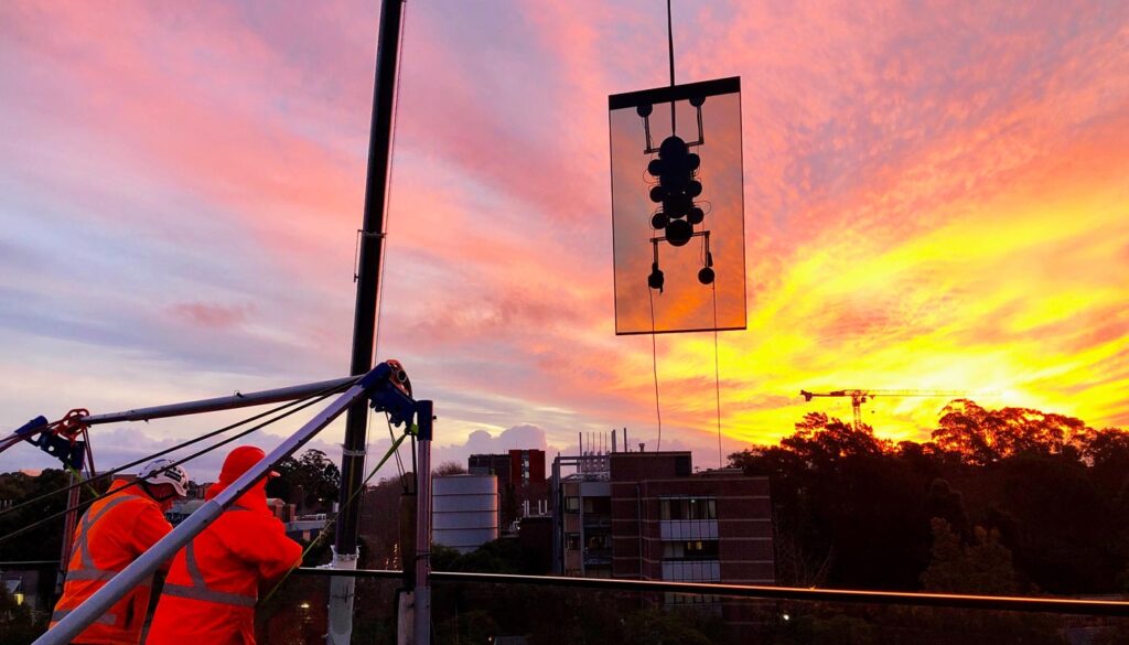 Construction workers lift a large pane of glass against a vibrant sunset sky.