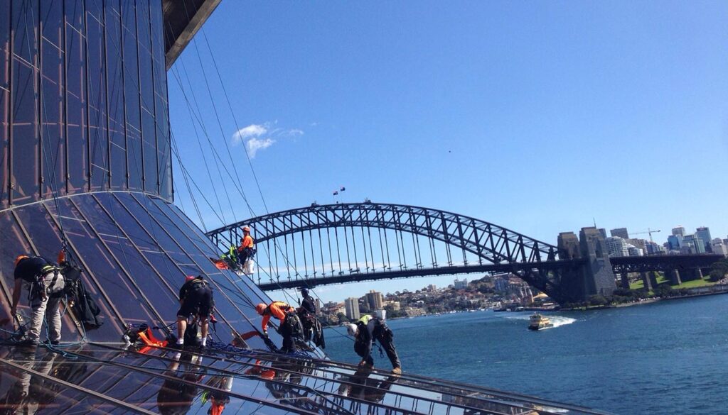 Window washers work on a glass building with the Sydney Harbour Bridge visible in the background.