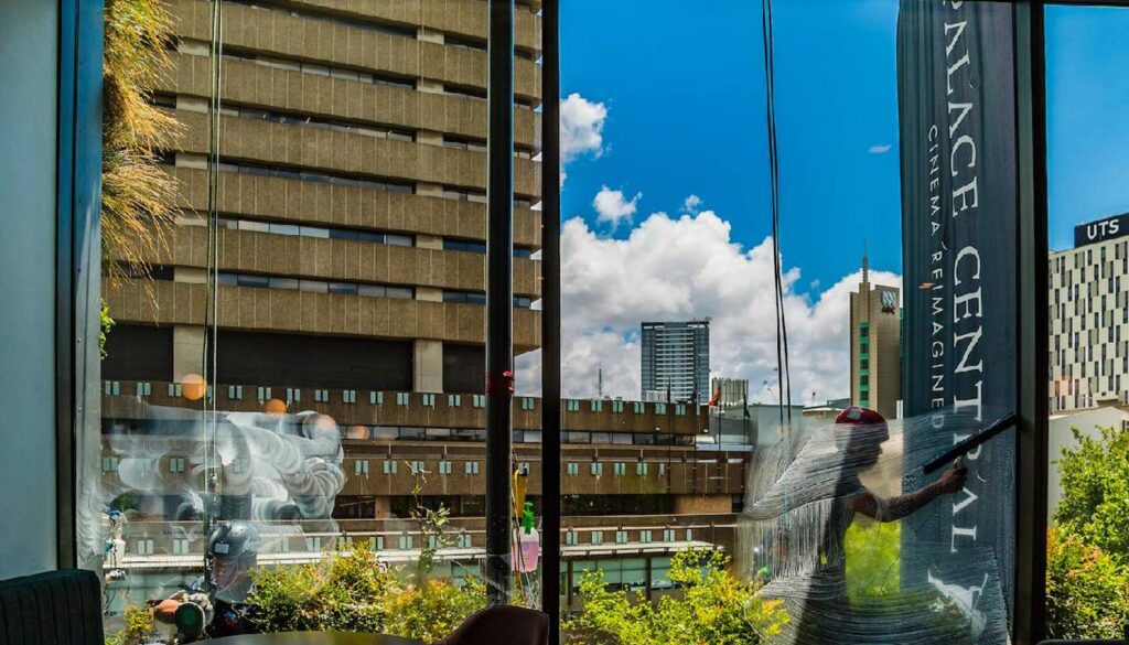A view through a window showing city buildings and a blue sky with clouds, framed by dark window mullions and reflected sculptures.
