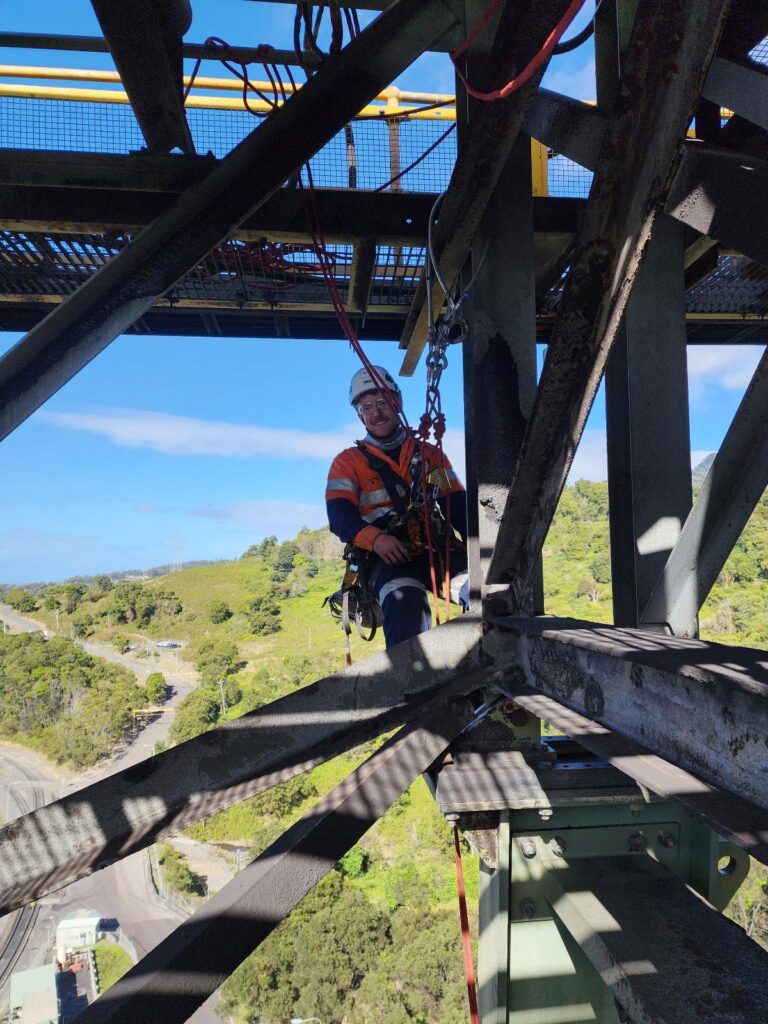 A worker wearing safety gear is suspended high on a metal structure, likely a tower or bridge, with a scenic landscape in the background.