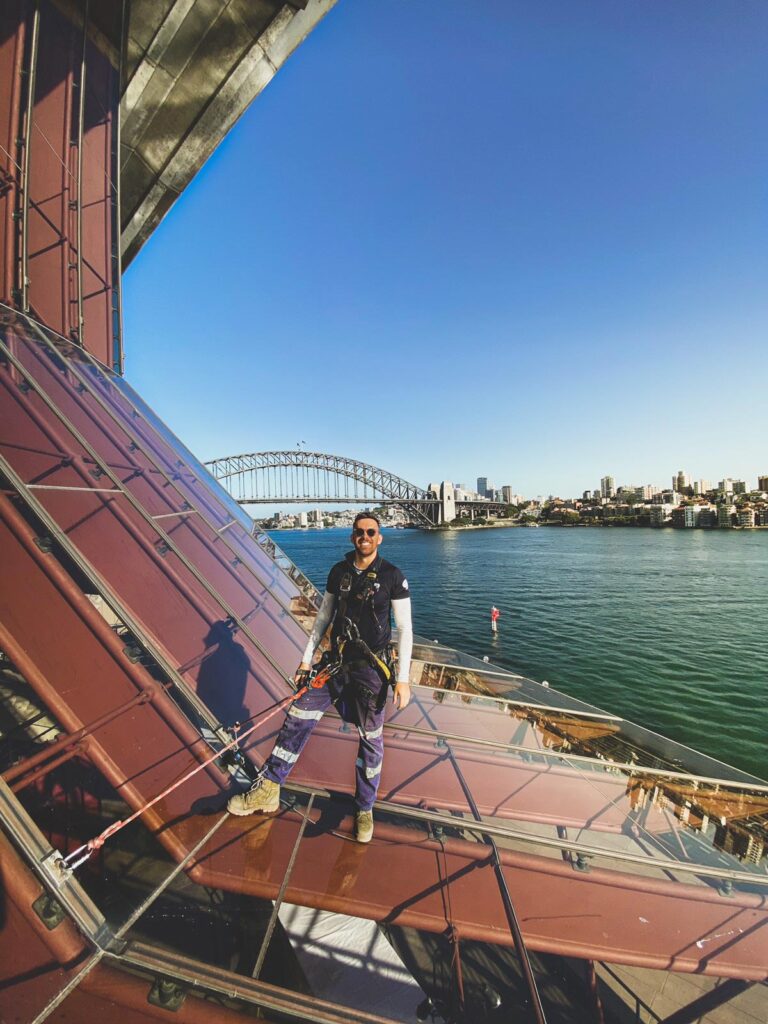 A window washer wearing safety gear stands on the exterior of a building with a view of the Sydney Opera House and Harbour Bridge.