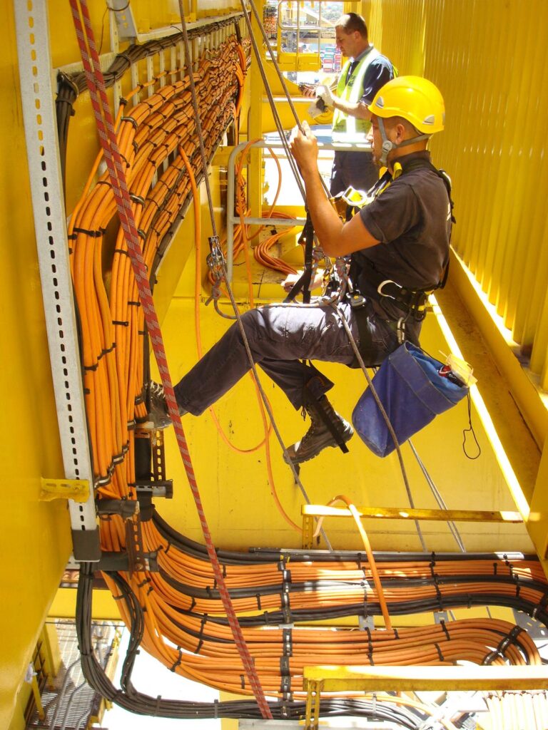 A worker wearing a harness and hard hat is suspended while working on cables inside a yellow structure.