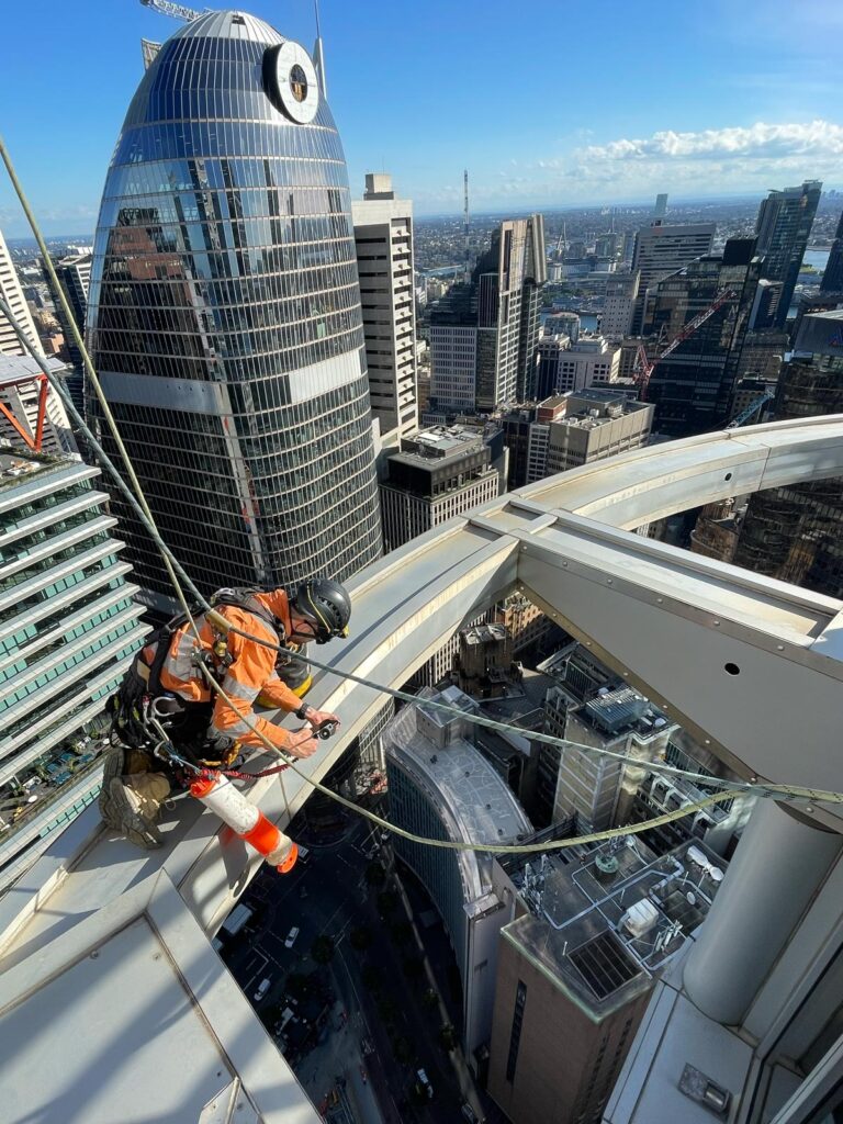 A window cleaner is suspended from a high-rise building, cleaning windows with a city skyline in the background.