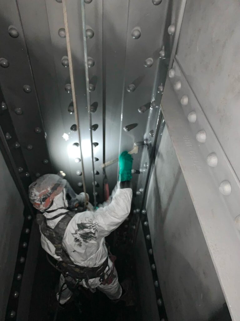 A worker in protective gear is rappelling down the inside of a large, metal industrial structure.