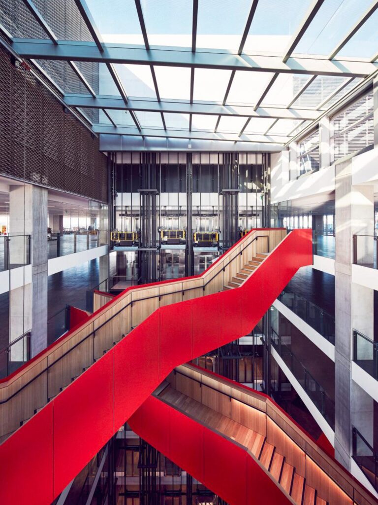 A modern architectural interior featuring a striking red staircase within a multi-level building with a glass ceiling.