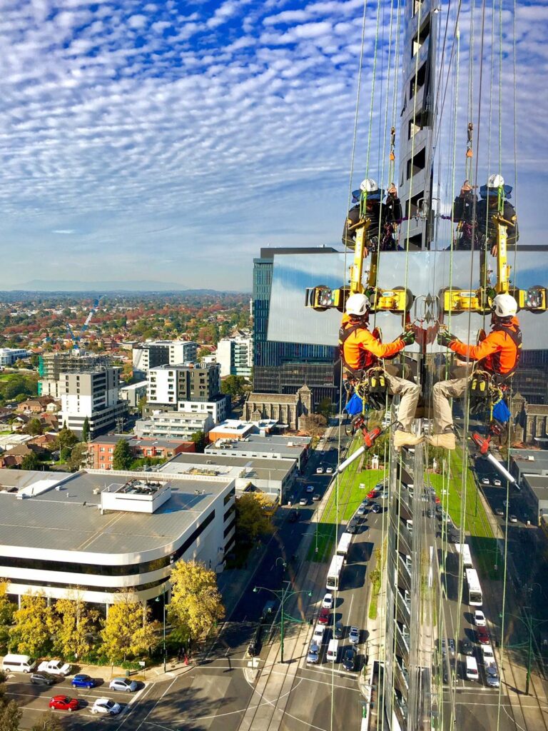 Construction workers are suspended on cables, installing glass panels on a high-rise building with a city skyline in the background.