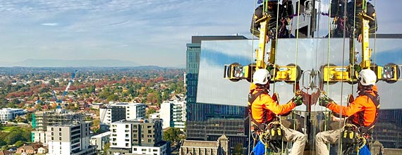 Two window washers in safety harnesses are cleaning the exterior of a tall skyscraper with a city view in the background.
