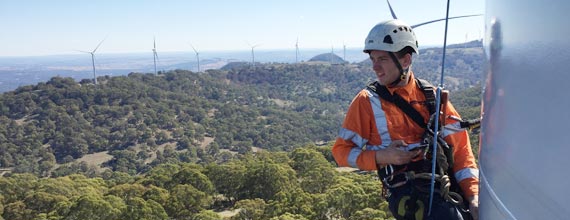 A worker wearing safety gear is rappelling down the side of a wind turbine with a scenic view of a wind farm and forested hills in the background.