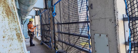A construction worker wearing a yellow hard hat and safety harness stands on a platform next to a building facade covered in safety netting.